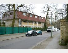 Wohnstraße mit einem zweistöckigen Wohngebäude, dessen Fassade teils aus gelbem Klinker besteht und über ein rotes Satteldach verfügt. Vor dem Gebäude stehen mehrere geparkte Autos, und der Bereich ist von Bäumen und einem grünen Zaun umgeben.