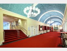 Großer, eleganter Eingangsbereich eines Veranstaltungsortes mit rotem Teppichboden, gewölbter blauer Decke, Kronleuchtern und Treppenstufen. An den Wänden befinden sich weiße Türen, Schaukästen und Bilder, während rote Vorhänge und Stehtische den Raum säumen.