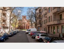 Straßenansicht in einer Stadt mit parkenden Autos auf beiden Seiten der Straße. Im Hintergrund steht eine Backsteinkirche mit spitzem Dach und großen gotischen Fenstern, umgeben von Bäumen.