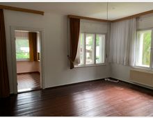 Wohnraum mit dunklem Holzboden, weißen Wänden und zwei großen Fenstern mit weißen Spitzenvorhängen und braunen Dekovorhängen. Im Hintergrund führt eine Türöffnung zu einem Raum mit rot-weiß kariertem Boden und einem weiteren Fenster mit braunen Vorhängen.