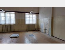 Leerer Raum mit hohen Decken, zwei großen Fenstern mit Gardinen, Heizkörpern unter den Fenstern und abgenutztem Bodenbelag. An einer Wand sind Kacheln und freiliegende Rohre sichtbar, während einige Paneele lose auf dem Boden liegen.