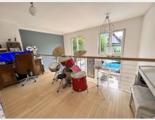 Offener Galerieraum in einem Wohnhaus mit hellem Holzboden, großem Fenster und Geländer mit Holzhandlauf über einer Treppe. Im Vordergrund steht ein rotes Schlagzeug, daneben hängen mehrere runde Lampenschirme an einer Pendelleuchte.