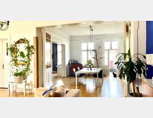 Heller Wohnraum in einem Altbau mit hohen Decken, weißen Wänden und Holzboden. Zu sehen sind ein großer Esstisch mit Stühlen, ein goldgerahmter Spiegel mit Pflanzen, sowie Fenster mit Blick nach draußen; eine Katze sitzt neben einer Balkontür.