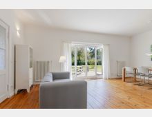 Heller Wohn- und Essbereich mit weiß gestrichenen Wänden und breiten Holzdielen, ausgestattet mit grauem Sofa, Vitrine und Esstisch mit Stühlen. Große verglaste Terrassentür mit weißen Vorhängen führt auf eine Terrasse und in einen grünen Garten.