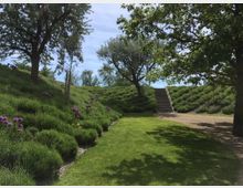 Ein parkähnlicher Garten mit einer Rasenfläche in der Mitte, umgeben von Lavendelbeeten und mehreren Bäumen. Im Hintergrund führt eine Treppe aus hellem Stein auf eine erhöhte Ebene, während der Himmel klar und blau ist.