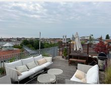 Dachterrasse mit Holzboden, ausgestattet mit weißen Sofas, kleinen runden Tischen, einem Sonnenschirm und einem Grill. Im Hintergrund ist eine städtische Skyline mit Gebäuden, Bäumen und bewölktem Himmel zu sehen.