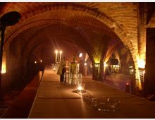Historischer Gewölbekeller mit Backsteinbögen, beleuchtet von Kerzen und warmem indirektem Licht. Ein langer, gedeckter Tisch mit Gläsern und Flaschen zieht sich durch den Raum.