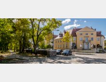 Straßenszene mit einem gelben Wohngebäude im klassischen Stil und roten Ziegeldächern, das von Bäumen und parkenden Autos umgeben ist. Im Vordergrund sind ein gepflasterter Gehweg, ein niedriger Steinwall und ein kleiner begrüner Parkbereich sichtbar.