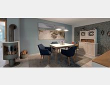 Esszimmer mit modernem Kaminofen links, einem rechteckigen Esstisch mit weißen Oberflächen und vier blauen Polsterstühlen. Im Hintergrund eine blaue Wand mit großem Landschaftsbild, rechts ein weißer, rustikaler Schrank.