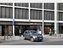 Fassade eines modernen Gebäudes mit einer Einfahrt zu einem Parkhaus, gekennzeichnet durch Schilder mit den Logos von „Q-Park“, „Fürst“ und „Rewe“. Rechts daneben befindet sich der Eingang zu einem „Rewe“-Supermarkt, während ein geparktes Auto und Fußgänger im Vordergrund sichtbar sind.