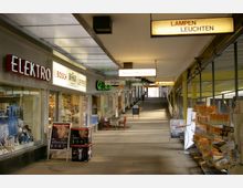 Überdachte Ladenpassage mit mehreren Geschäften, darunter ein Elektrofachgeschäft und ein Restaurant, sowie beleuchteten Schildern wie "Lampen Leuchten" und "Siemens Hausgeräte". Die Passage hat eine leicht ansteigende Rampe und Geländer auf der rechten Seite.