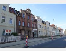 Straßenansicht mit einer Reihe von mehrstöckigen Wohnhäusern, darunter ein renovierungsbedürftiges Gebäude mit bröckelnder Fassade. Auf dem Bürgersteig befinden sich Bauarbeiten, gekennzeichnet durch rot-weiße Absperrungen und Warnlichter.
