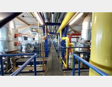 Industrielle Anlage mit einem erhöhten Gang aus Metallgittern, umgeben von blauen Geländern, Rohrleitungen in Gelb und Rot sowie großen Metallbehältern. Die Decke ist mit Metallverkleidung und sichtbaren Dachbalken gestaltet, und es verlaufen zahlreiche Lüftungsrohre.