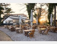 Outdoor café patio with many wooden tables and folding chairs on a gravel surface, dotted with tall closed umbrellas. A low white building with large windows sits behind the seating area, surrounded by tall trees with sunlight filtering through.