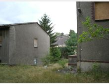 Zwei verfallene Gebäude mit grauem Putz, teilweise mit Brettern vernagelten Fenstern, stehen inmitten von hohem Gras und vereinzelten Büschen. Im Hintergrund ist ein weiteres Gebäude mit einem beschädigten Dach sowie Bäume und dichte Vegetation zu sehen.