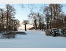 Schneebedeckter Park mit kahlen Bäumen, einer Statue in einem Brunnen im Vordergrund und einer kleinen Villa im Hintergrund. Im Hintergrund ist ein See sichtbar, eingefasst von weiteren Bäumen unter klarem Himmel.