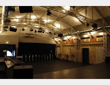 Innenansicht eines Veranstaltungsraums mit industriellen Elementen, wie einer Backsteinwand, freiliegenden Lüftungsrohren und Stützträgern aus Metall. Der Raum verfügt über eine Bühne mit Scheinwerfern an einer Deckenkonstruktion und schwarzen Vorhängen sowie eine offene Fläche mit wenigen Tischen und Stühlen.