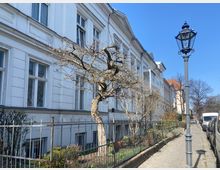 Eine ruhige Straßenansicht mit einer Reihe eleganter, weißer Altbauten im klassischen Stil. Im Vordergrund ein kahler Baum hinter einem schmiedeeisernen Zaun, daneben ein altertümlicher Laternenpfahl; am rechten Straßenrand parken Autos.