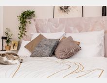 Blick auf ein gemütliches Schlafzimmer mit hellem Bett, dekoriert mit mehreren Kissen in unterschiedlichen Texturen und Mustern sowie einer gemusterten Tagesdecke. Im Hintergrund befindet sich ein rosa gemusterter Bettrücken, an der Wand hängen Kunstwerke, und links steht ein Nachttisch mit einer Pflanze und einer Lampe.