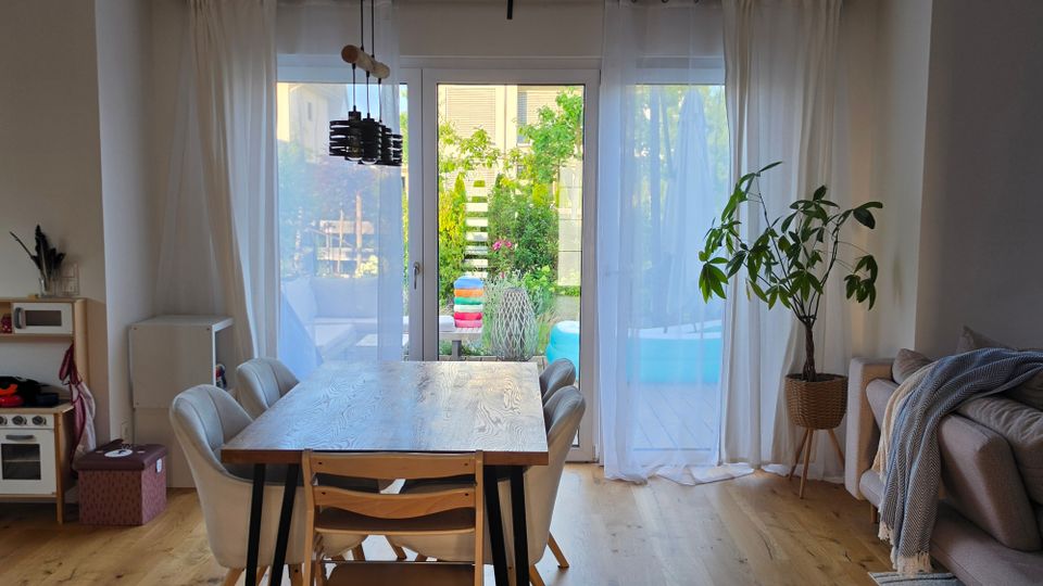 Heller Wohn- und Essbereich mit Holzboden, Esstisch und Stühlen sowie großer Glas-Schiebetür mit weißen Vorhängen zur Terrasse und in den Garten. Rechts stehen eine Topfpflanze und ein Sofa, links ist eine kleine Küchen- bzw. Spielküchenecke zu sehen.