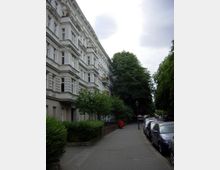 Straßenszene mit historischen Wohngebäuden auf der linken Seite, gekennzeichnet durch weiße Fassade, klassische Stuckelemente und Balkone. Rechts säumen geparkte Autos und hohe Bäume den gepflasterten Gehweg.