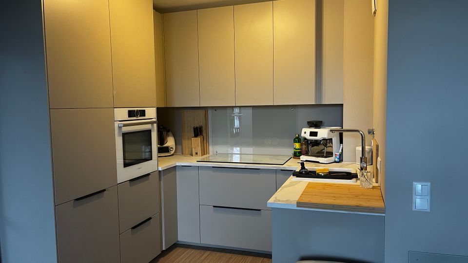 Moderne, kompakte Einbauküche in L-Form mit hellen, grifflosen Schränken und integrierten Geräten wie Backofen und Kochfeld. Auf der Arbeitsplatte stehen Spüle, Kaffeemaschine und Küchenutensilien; der Boden ist in Holzoptik und die Beleuchtung erfolgt über eine Deckenleuchte.