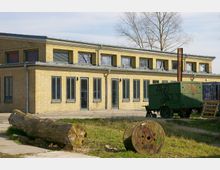 Ein einstöckiges Gebäude aus gelben Ziegelsteinen mit großen grauen Fenstern und einem flachen Dach; im Vordergrund befinden sich ein Baumstamm, eine bemalte Holzspule und ein grüner Anhänger mit Graffiti. Im Hintergrund stehen kahle Bäume vor einem hellen Himmel.