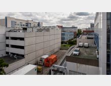 Mehrstöckiges Parkhaus und umliegende Bürogebäude in einem urbanen Gewerbegebiet. Im Vordergrund sind geparkte Fahrzeuge, darunter Transporter auf einer Rampe und Container neben einem Kundeneingang, sichtbar.