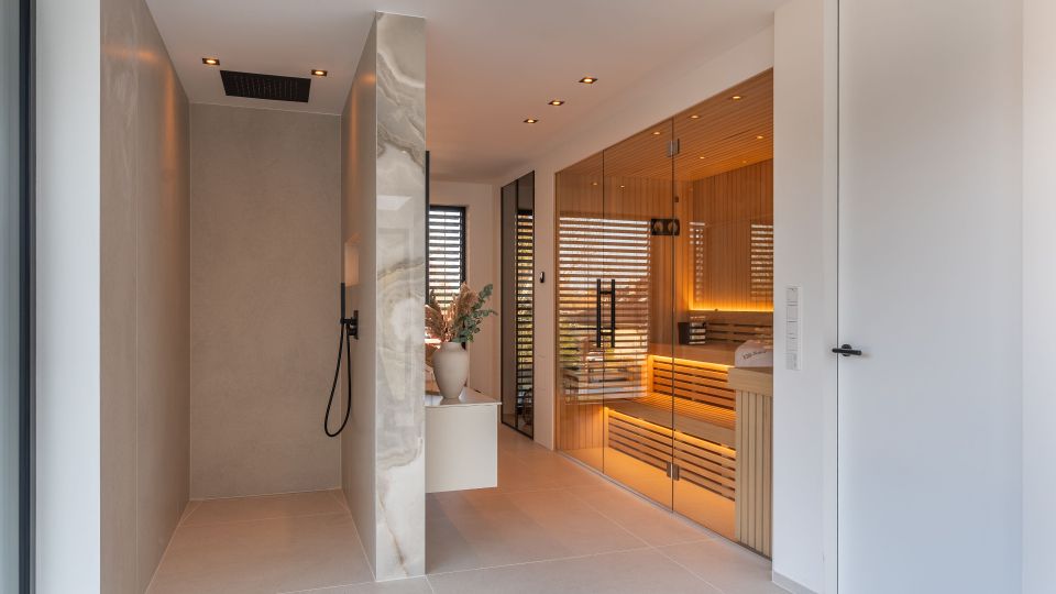 Moderner Wellnessbereich mit großformatigen Fliesen, bodengleicher Dusche mit schwarzer Armatur und einer verglasten Sauna aus Holz mit indirekter Beleuchtung. Schmale Fenster mit Lamellen, Einbauspots in der Decke und eine freistehende Trennwand aus Stein strukturieren den Raum.