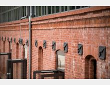 Fassade eines Gebäudes aus rotem Backstein mit kleinen, gleichmäßig verteilten Lüftungsgittern. Im unteren Bereich befinden sich bogenförmige Fenster und Türen mit modernen Glaselementen.