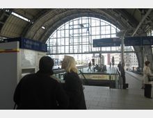 Ein moderner S-Bahnhof mit gewölbtem Glasdach und großen Fensterflächen, durch die Tageslicht einfällt. Im Vordergrund stehen mehrere Personen an einem Fahrkartenautomaten, während im Hintergrund Gleise und ein Bahnsteig mit wartenden Reisenden sichtbar sind.