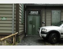 Außenansicht eines Gebäudes mit grün gestrichenen Holztüren und Fensterläden, daneben ein Schild mit der Aufschrift „Eingang zum Restaurant“ und ein geparktes silbernes Auto auf einer mit Pflastersteinen bedeckten Fläche. Die Fassade besteht aus holzverkleideten Wänden in braun-grüner Optik.