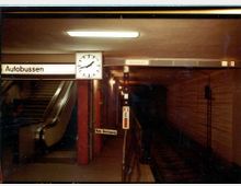 U-Bahn-Station mit einer Wanduhr, einem Schild mit der Aufschrift „Autobussen“ und einem Rolltreppenbereich links. Rechts ist ein dunkler Tunnel zu sehen, daneben Schilder mit Fahrtrichtungsangaben.
