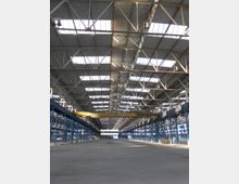 Weitläufige Industriehalle mit modularer Stahlkonstruktion, blauen Tragwerken und einem hohen Dach mit mehreren großen Oberlichtern. Über der Halle hängt ein gelber Laufkran, der entlang von Schienen an den Seitenwänden montiert ist.