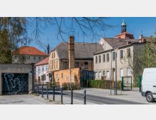 Straßenszene in einem städtischen Wohngebiet mit älteren Gebäuden, darunter ein verputztes Haus mit einem Schornstein und mehrere beige Wohnhäuser. Im Hintergrund sind der Turm einer Kirche sowie rote Ziegeldächer zu sehen, während ein Gehweg, eine Straße und ein geparktes weißes Fahrzeug im Vordergrund stehen.
