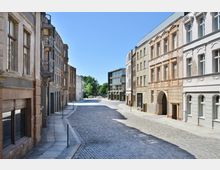 Eine gepflasterte Straße in einem historischen Viertel mit mehrstöckigen Gebäuden aus hellem und dunklem Putz, einigen mit Balkonen. Im Hintergrund sind moderne Gebäudeelemente und grüne Bäume zu sehen, unter einem klaren, blauen Himmel.