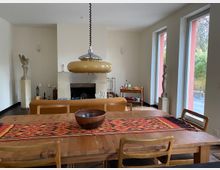 Essbereich in einem hellen Wohnzimmer mit großem Holztisch und Stühlen, gemustertem Tischläufer und hängender Deckenlampe. Im Hintergrund sind ein Kamin, Skulpturen und ein Beistelltisch zu sehen; rechts sorgen hohe Fenster mit Blick ins Grüne für viel Tageslicht.