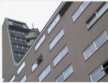 Ein modernes Gebäude mit glatter Fassade und großen, symmetrisch platzierten Fenstern, das sich um einen schrägen, höher gebauten Turm mit abgerundeten Balkonen und einer Metallstruktur erweitert. Der Himmel ist wolkenbedeckt und bildet den Hintergrund.