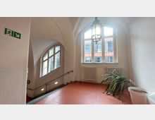 Treppenhaus mit hohen, weiß gestrichenen Wänden und großen, bogenförmigen Fenstern, durch die Tageslicht fällt. Links ist ein Notausgangsschild zu sehen, rechts eine Topfpflanze vor einem Heizkörper unter den Fenstern.