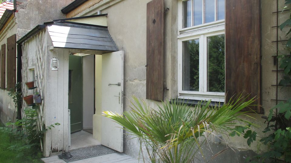 Eingangsbereich eines älteren, verputzten Hauses mit kleiner überdachter Holzveranda und offener Tür. Daneben ein Fenster mit hölzernen Klappläden; im Vordergrund stehen eine Topfpalme und etwas Gartenbewuchs.
