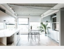 Moderne Küche mit minimalistischem Design, einem hohen weißen Tisch mit schwarzen Hockern, Edelstahlarbeitsflächen und einem freiliegenden Beton-Deckenstil. Große Fenster lassen viel Tageslicht herein, und eine Palme sowie eine kleine Pflanze ergänzen die Einrichtung.