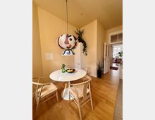 Essbereich in einem hellen Raum mit Holzfußboden, einem runden weißen Tisch, vier Holzstühlen mit gewebten Sitzflächen und einer bunten, dekorativen Hängelampe. Im Hintergrund stehen Pflanzen, ein hoher Schrank und ein geöffneter Durchgang zu einem weiteren Raum.