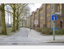 Wohnstraße mit mehrstöckigem Gebäude aus braunem Putz und gelb umrahmten Fenstern auf der rechten Seite, einem Gehweg und Bäumen auf der linken Seite. Verkehrsschilder kennzeichnen eine Tempo-30-Zone und eine Sackgasse.