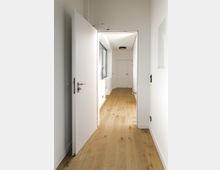 Korridor in einem modernen Innenraum mit hellen Wänden, einer offenen weißen Tür und einem Boden aus hellem Holz. Links befindet sich ein Fenster mit Jalousien; am Ende des Flurs ist eine geschlossene weiße Tür sichtbar.