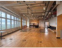 Large open-plan studio or office space with a light wood floor, floor-to-ceiling windows along one side, and an exposed industrial ceiling with visible ducts, pipes, and hanging lights. A small kitchenette/bar area sits at the far end, with high stools by the windows and a long bench against the wall.