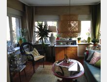 Wohnzimmer mit Holzboden, vielen Pflanzen und einer großen Fensterfront, die von Vorhängen umrahmt ist. Es gibt eine Sitzgruppe mit einem geflochtenen Sessel, einem Sofa mit bunten Kissen und einem Holztisch mit Glasplatte. Eine hängende Rattanlampe und dekorative Gegenstände, wie Vasen und Teekannen, verleihen dem Raum eine gemütliche Atmosphäre.