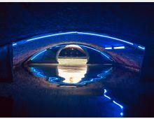 Unterirdischer Tunnel mit gewölbter Backsteinarchitektur, einer Wasseroberfläche im Vordergrund und blauen LED-Lichtstreifen, die die Decke und Wände akzentuieren. Im Hintergrund erhellt ein beleuchteter Durchgang die Szene.