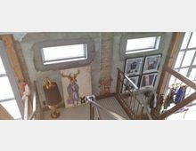 Treppenhaus in einem modern gestalteten Innenraum mit Betonwänden, sichtbaren Holzbalken und großen Fenstern. An den Wänden hängen Kunstwerke, darunter ein großes Bild mit einer Tierfigur, sowie gerahmte Illustrationen; daneben befinden sich ein goldfarbener Lampenfuß in Tierform und eine Kleiderstange mit Kleidung.