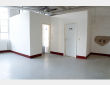 Innenraum einer industriellen Halle mit glattem grauem Boden, hohen Fenstern und sichtbaren Deckenrohren. In der Mitte steht ein weißer, boxförmiger Einbau mit zwei Türen, eine davon mit „WC“-Schild, sowie rotem Sockelabschluss.