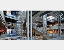 Modernes Bahnhofsinterieur mit mehreren Ebenen, transparenten Aufzügen aus Glas und einer breiten Rolltreppe. Im Hintergrund befinden sich Geschäfte und Werbetafeln, umgeben von einer Stahl- und Glasarchitektur.